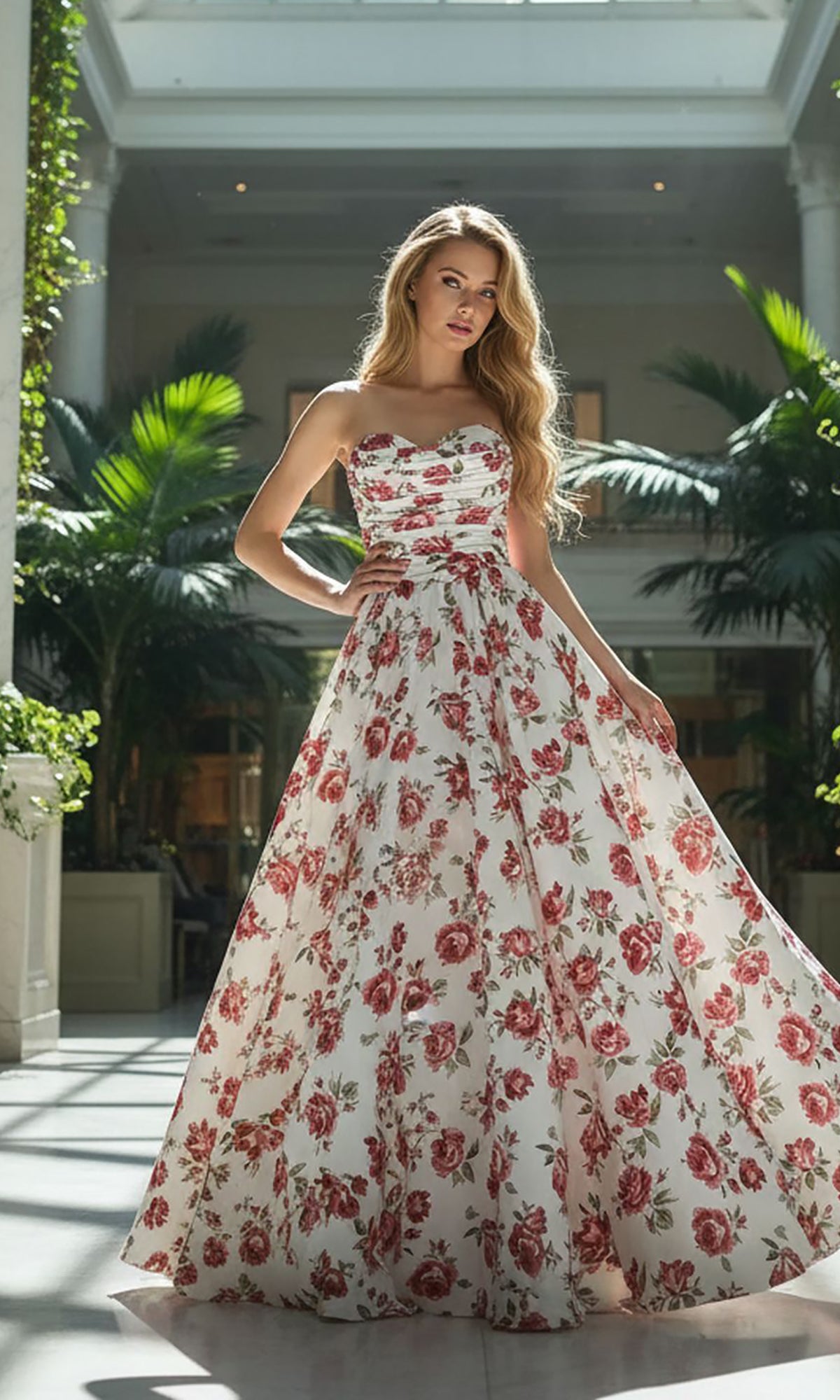 A woman stands indoors in the Amelia Couture 8070 long prom dress in white. Lush green plants and tall columns in the background enhance her elegant appearance.